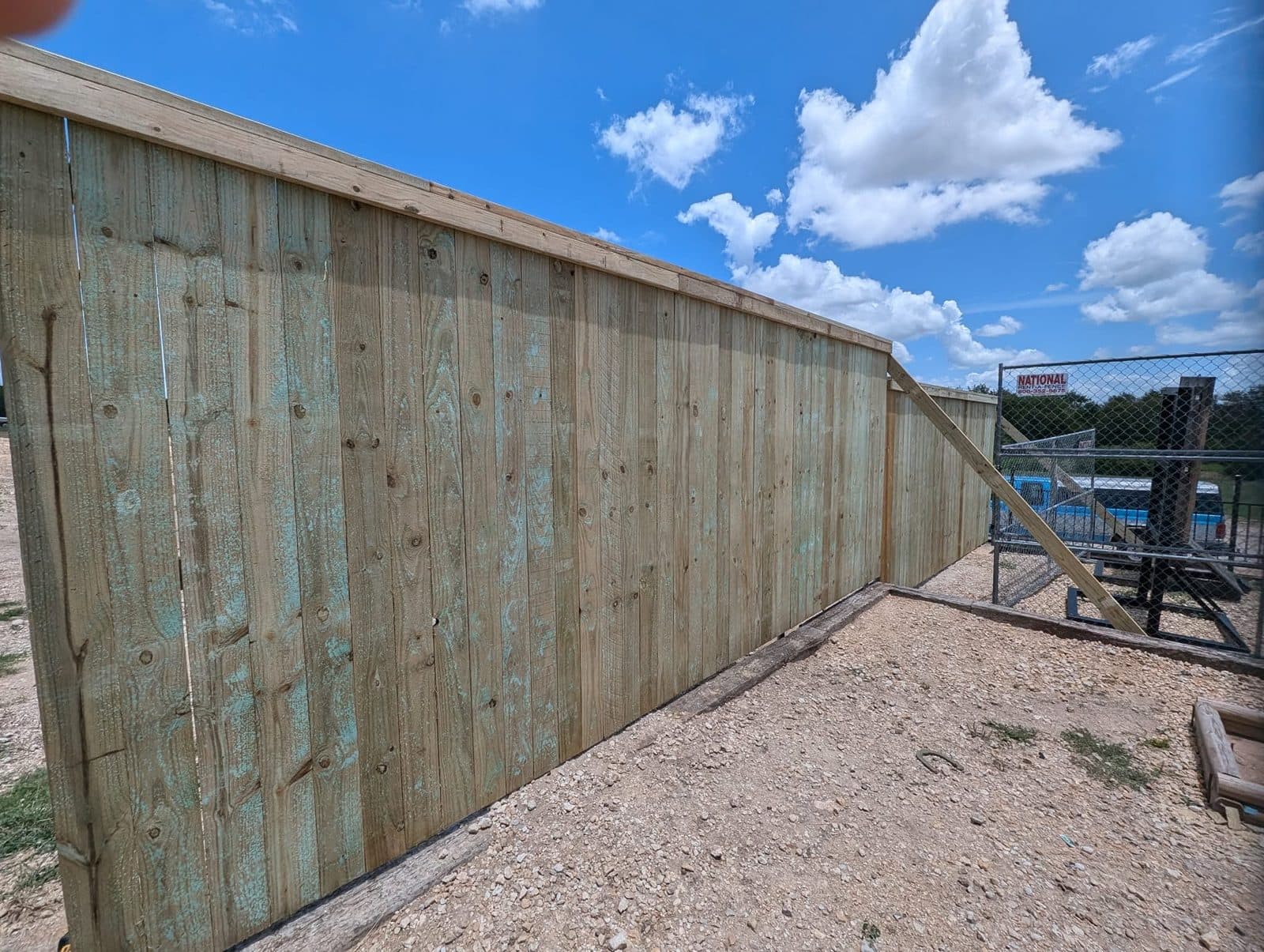 Finished face of a tall pressure-treated wood privacy fence with a cap rail, running along a gravel yard